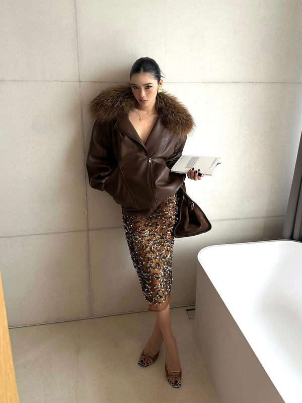 Woman in a stylish outfit with a fur-lined coat and sequin skirt, leaning against a wall.