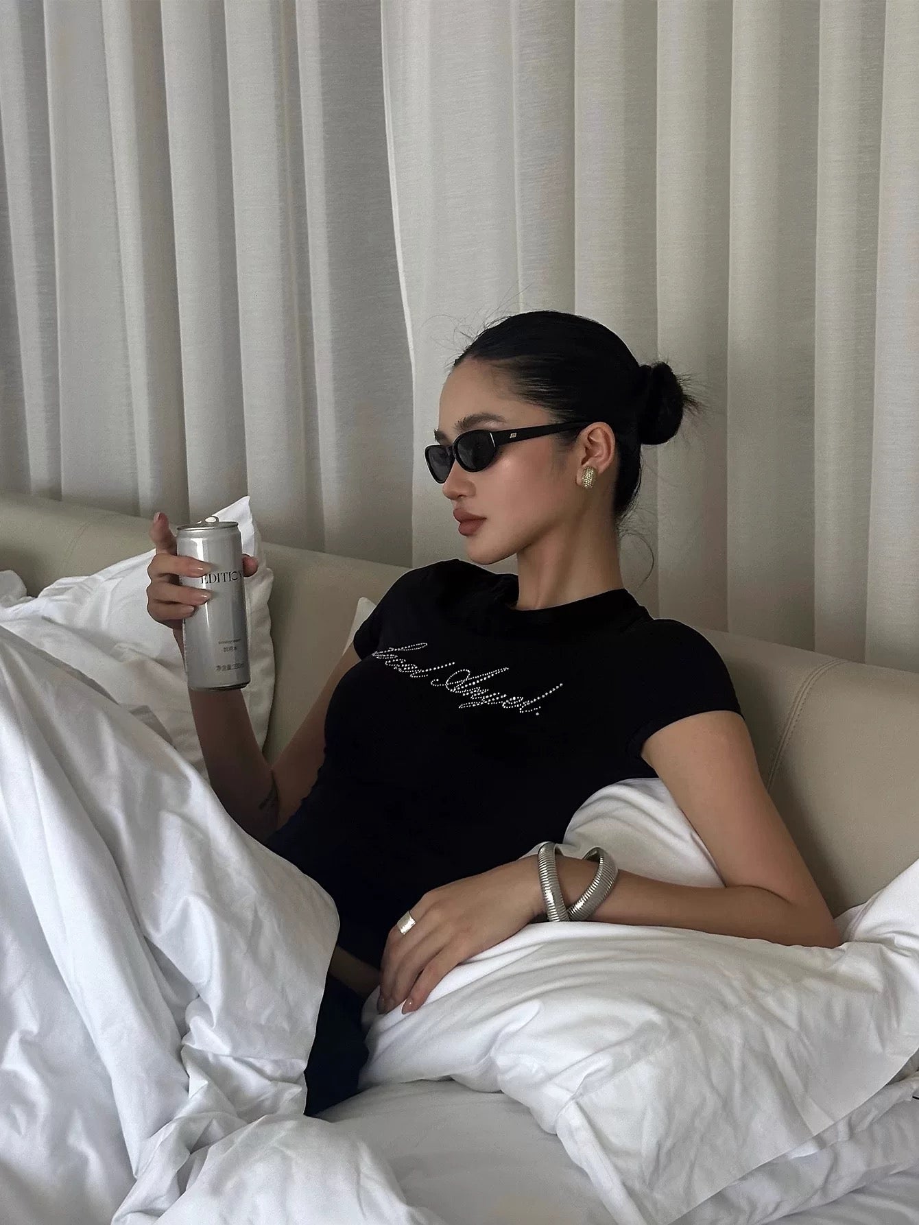 Woman sitting on a bed holding a drink, wearing sunglasses and a black shirt with local angel text on the t-shirt..