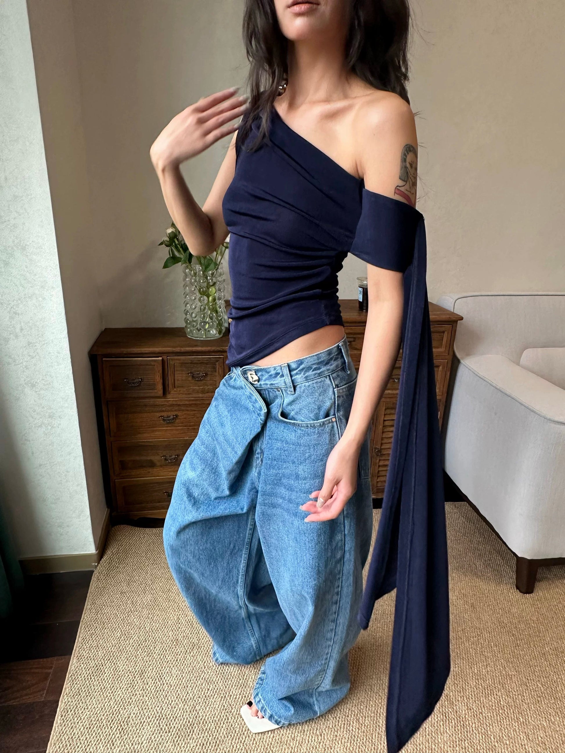 Person wearing a navy one-shoulder top and blue jeans indoors.