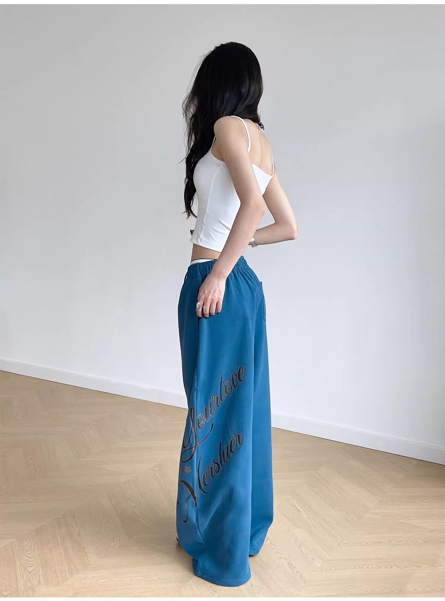 Woman wearing blue pants with text and a white top in a minimalistic room.