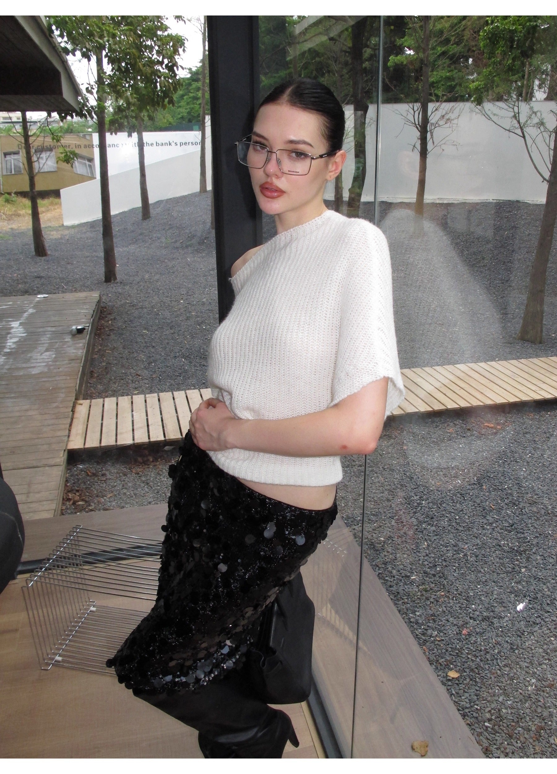 Woman wearing a white off the shoulder sweater with a black sequin skirt standing outdoors.