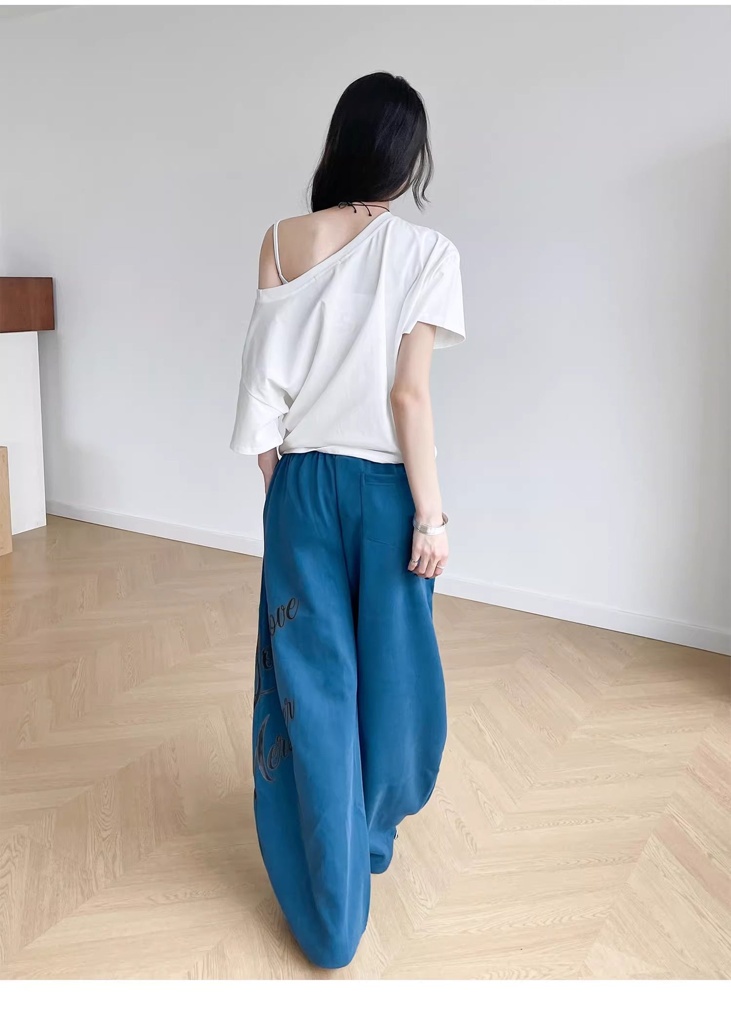 Person wearing a white top and blue pants standing in a minimalistic room with wooden flooring.
